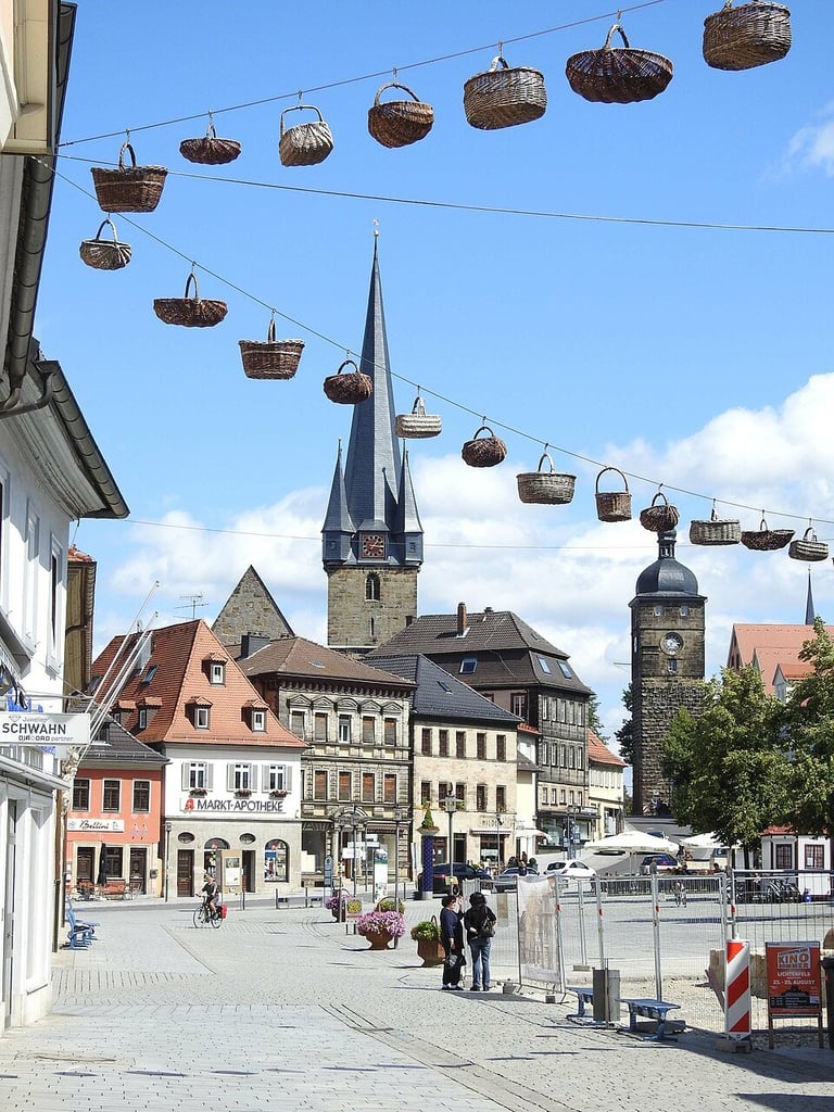 Historic European town square with church spire, colorful buildings, and decorative woven baskets hanging from overhead cables